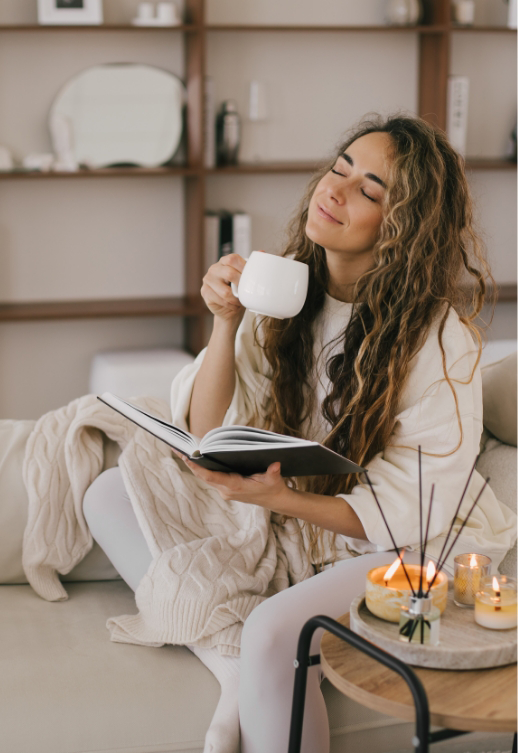 Frau entspannt auf einem Sofa, liest ein Buch und hält eine Tasse, im Hintergrund ein Regal.