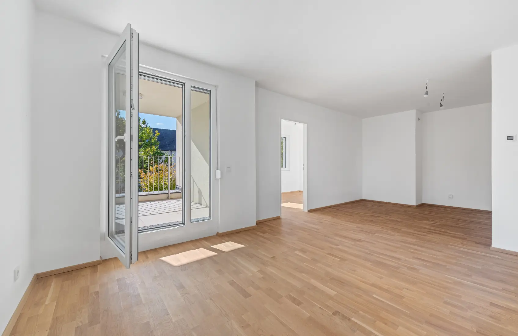 Heller, leerer Wohnraum mit Parkettboden und großen Glastüren, die zu einem Balkon führen.
