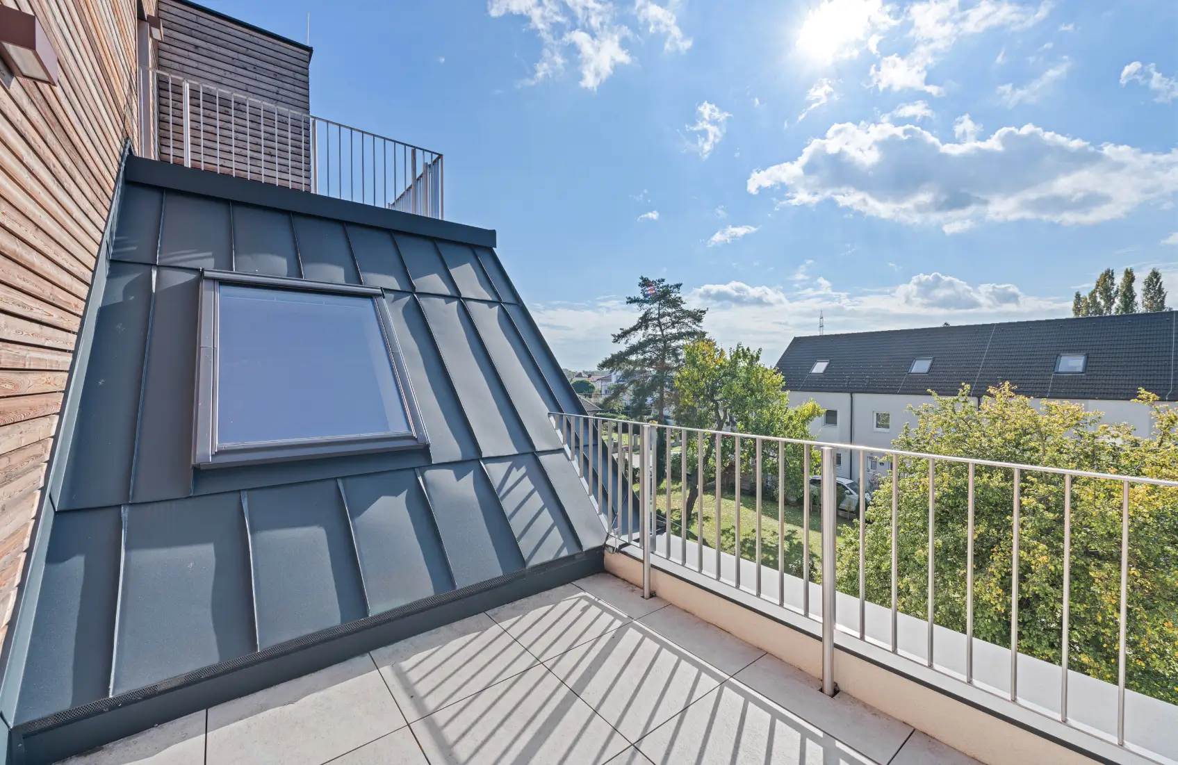 Balkon oder Terrasse mit Metallgeländer, Dachschräge und Blick über Wohnhäuser und Bäume.